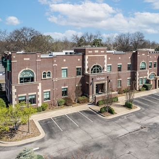 Brick office building with arched windows surrounded by trees and empty parking spaces under a blue sky