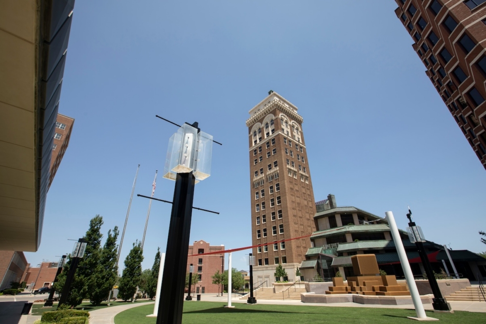 Tall brick building with clock tower and modern sculptures in an urban park setting under clear blue sky