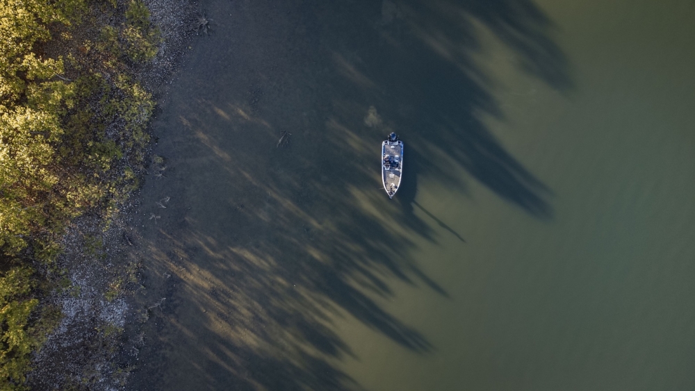 Aerial view of a boat on a calm lake near a wooded shoreline with long shadows.