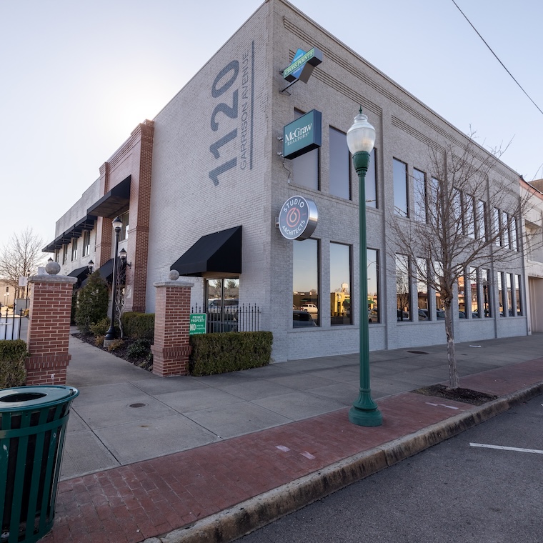 Two-story brick building with multiple signs and sidewalk, featuring trees and a green lamp post.