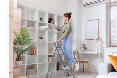 Woman cleaning tall white shelves in a modern living room using a step ladder