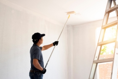 Man painting a ceiling with a roller near a ladder in a bright room